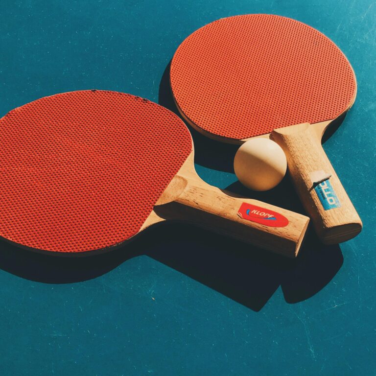 Two table tennis rackets and a ball on a blue background with sunlight casting shadows.